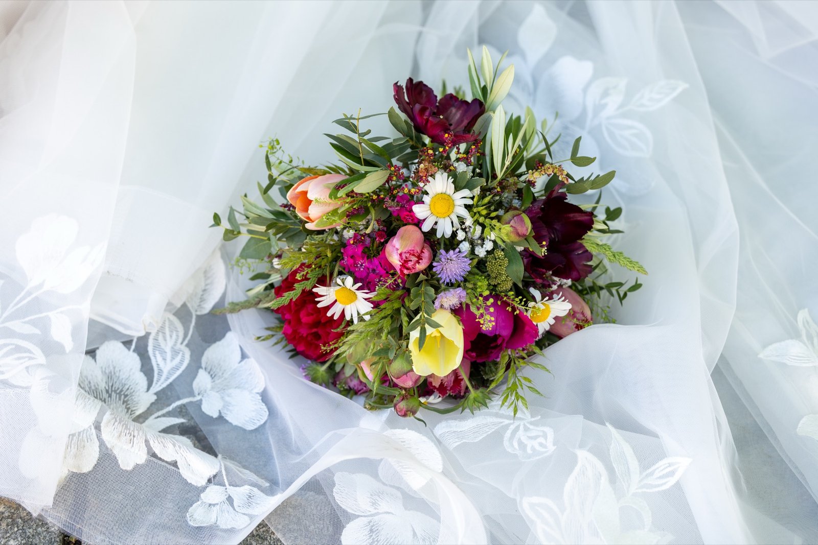 Ramo de novia colorido con tulipanes, margaritas y peonías sobre un velo blanco bordado